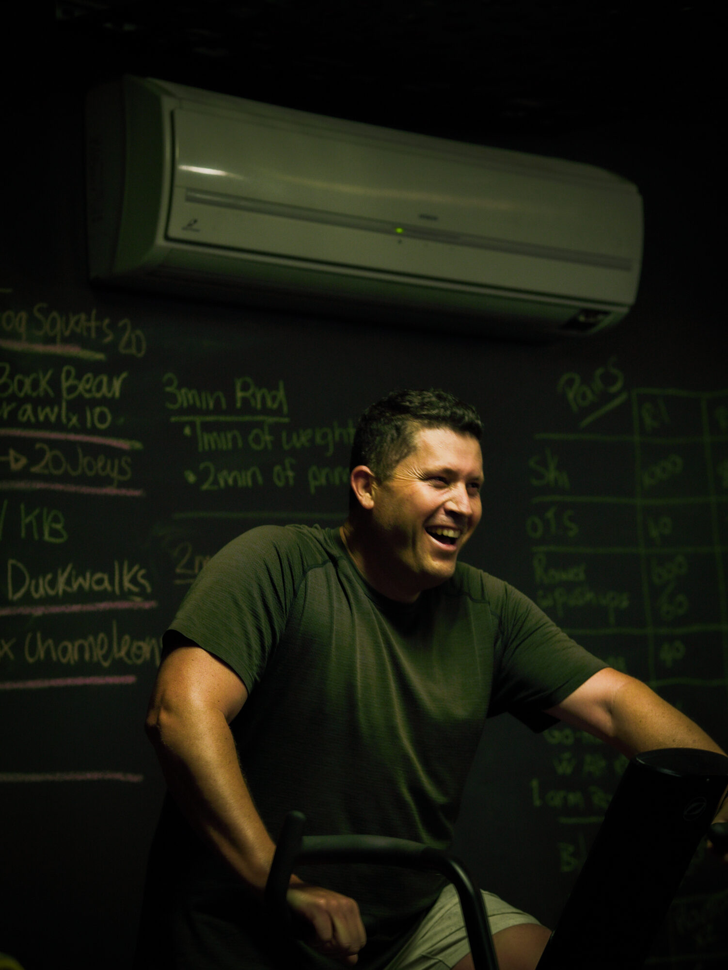 A member laughing on the air bike with the session board chalked up behind him