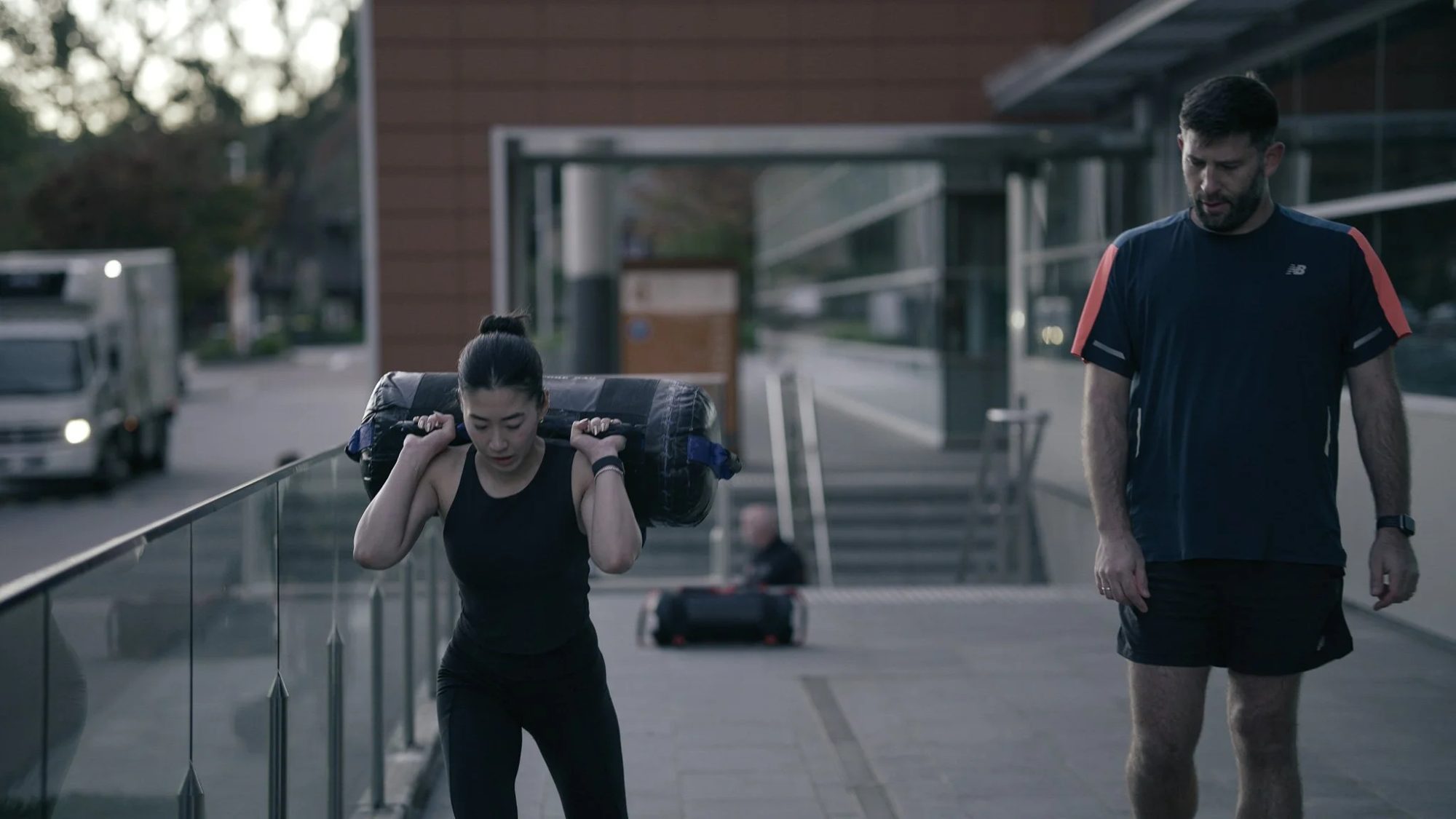 A coach walking beside a member carrying a sandbag outside the building