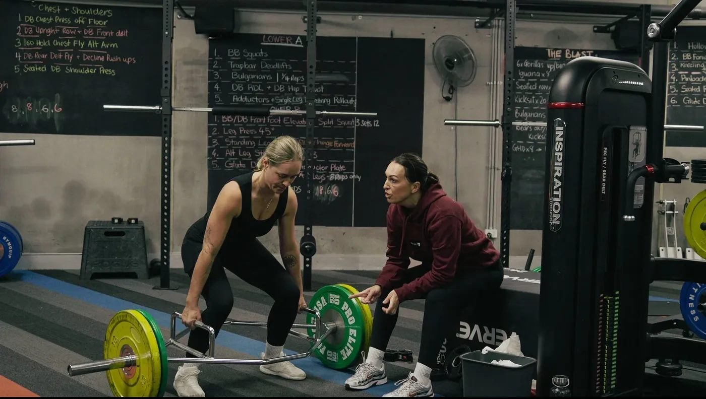 Nicole coaching a member through a trap-bar deadlift in the weights area