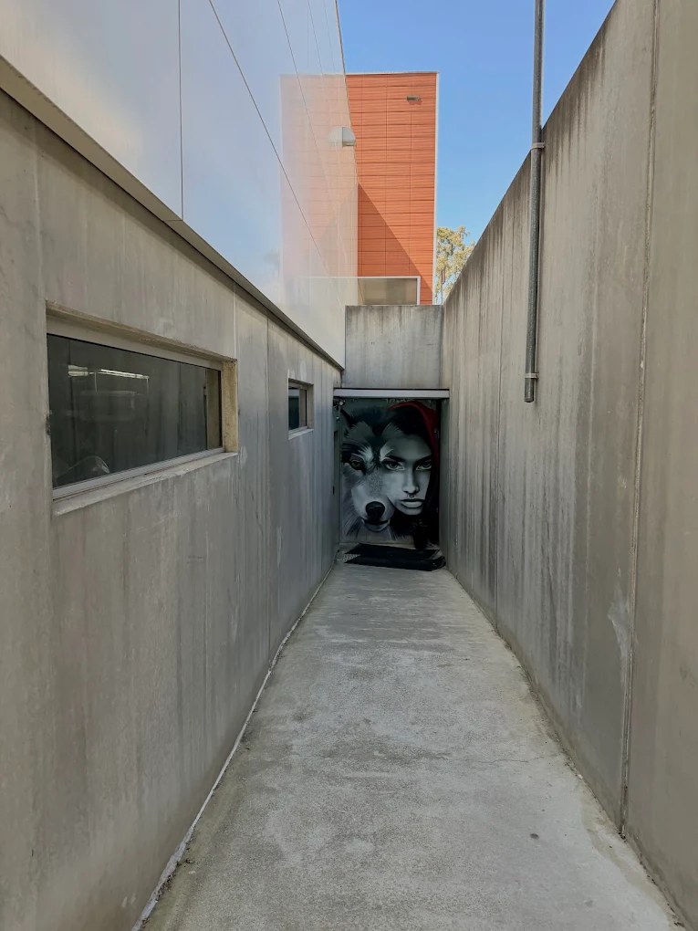 A narrow concrete walkway leading to a mural of a wolf and a woman in a red hood at The Den's entrance